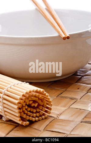 ein vertikales Bild von einem Paar Stäbchen auf eine weiße Schüssel mit einer Suhi Rollen Matte auf ein Platzdeckchen gewebt Stockfoto