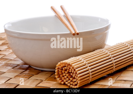 ein horizontales Bild von einem Paar Stäbchen auf eine weiße Schüssel mit einer Suhi Rollen Matte auf ein Platzdeckchen gewebt Stockfoto