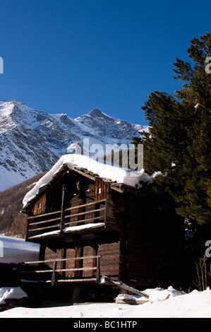 Traditionelle hölzerne Berghütte im Winter mit schneebedeckten Gipfeln und immergrünen Bäumen unter klarem blauem Himmel. Stockfoto