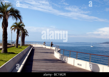 Sonnige Strandpromenade mit Palmen, Bänken und Spaziergänger mit Blick auf das ruhige blaue Wasser und die ferne Küste an einem klaren Tag Stockfoto
