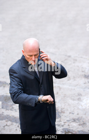 Glatzkopf in seinen späten 40er Jahren trägt einen dunklen Anzug sprechen auf einem Handy und mit Blick auf eine Armbanduhr. Stockfoto