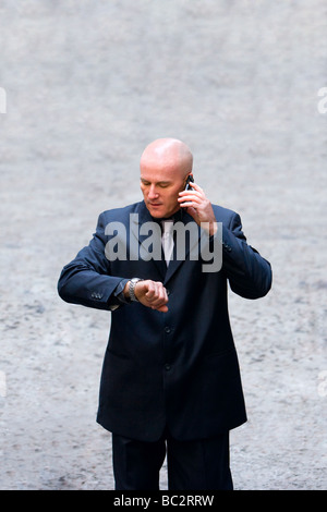 Glatzkopf in seinen späten 40er Jahren trägt einen dunklen Anzug sprechen auf einem Handy und mit Blick auf eine Armbanduhr. Stockfoto