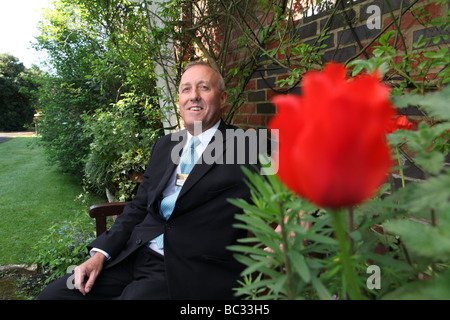 ein Porträt von einem Butler in einem Garten von einem stattlichen Haus lächelnd sitzt und schaut in die Kamera mit einer roten Rose im Vordergrund Stockfoto