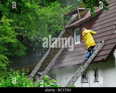 Mann trägt einen gelben Regenmantel stehen auf einer Leiter Reinigung Dach mit einem Hochdruck-Wasserschlauch Stockfoto