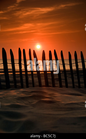 Die untergehende Sonne, die durch einen Zaun auf einem sandigen Strand gesehen Stockfoto