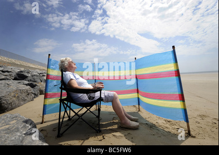 EIN RENTNER GENIEßT SONNENBAD AN EINEM BRITISCHEN STRAND MIT WINDBREAKER RE URLAUB RUHESTAND RENTNER RENTNER SONNE GEFAHR UK Stockfoto