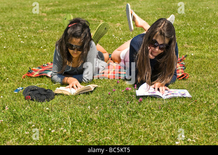 Horizontale Porträt von zwei attraktive junge Mädchen liegen auf dem Rasen, Bücher lesen und hören von einem Ipod an einem sonnigen Tag Stockfoto