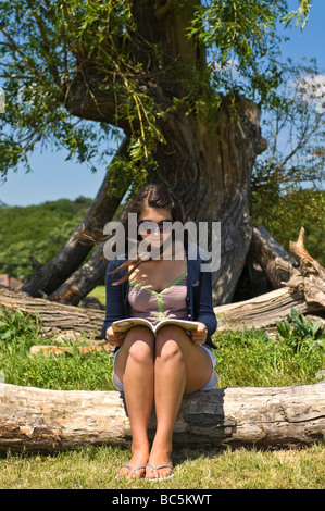 Vertikale Porträt eines attraktiven jungen Teenager Mädchen sitzen auf einem Baumstamm ein Buch unter einem Baum an einem sonnigen Tag Stockfoto