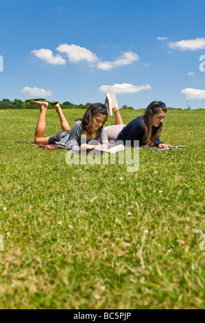 Vertikale Porträt von zwei attraktiven jungen Mädchen im Teenageralter liegen auf dem Rasen, Bücher lesen und hören von einem Ipod an einem sonnigen Tag Stockfoto