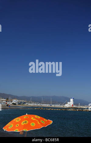 Eine Orange gefärbte Strand Sonnenschirm im Vordergrund mit Blick auf eine spanische Küste mit einem Leuchtturm und Yachten. Stockfoto