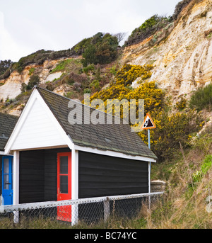 Strandhütten an der Basis der erodierenden Klippen. Ein Zeichen hinter den Hütten warnt vor der Gefahr von Steinschlag. Bournemouth direkt am Meer. Stockfoto