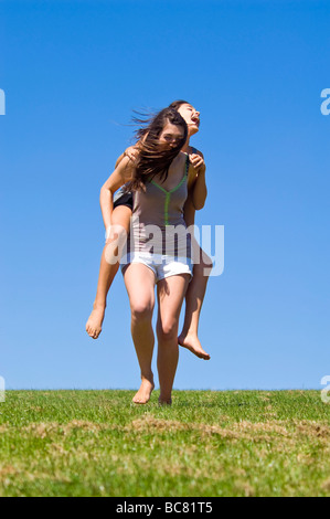 Vertikale Porträt von zwei jungen Schwestern, die mit viel Spaß mit Huckepack reitet auf einem sonnigen Tag Stockfoto
