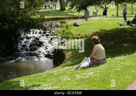 Single-Frau sitzen auf dem Rasen neben kleinen Wasserfall Buxton Pavilion Gardens im englischen Derbyshire Peak District Stockfoto