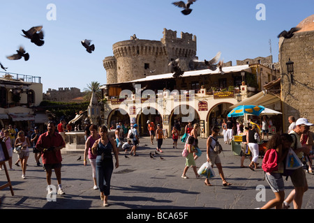 Menschen Flanieren über beschäftigt Platia Ippokratou mit Geschäften und Restaurants, fliegende Tauben, Thalassini Tor im Hintergrund, Rhodos Tow Stockfoto