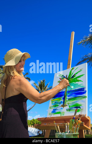 Einer weiblichen Künstlerin in den Fünfzigern erstellen ein Gemälde auf Leinwand von einer Palme und dem Ozean im Lanikai, Hawaii Stockfoto
