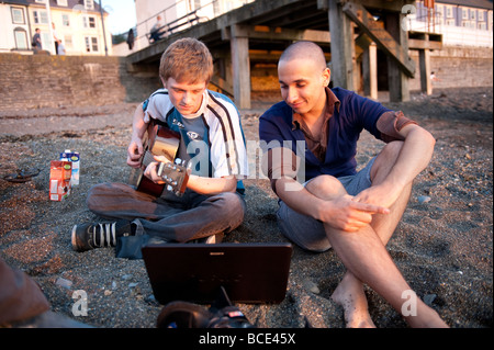 Ein junger Mann, Blick auf Laptop-Computer am Strand, während sein Freund auf Aberystwyth Wales UK sieht Gitarre spielen Stockfoto