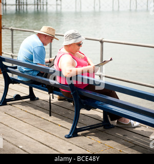 Ein Elederly paar sitzt auf einer Bank und am Meer liest Zeitschriften an einem sonnigen Sommertag am Meer UK Stockfoto