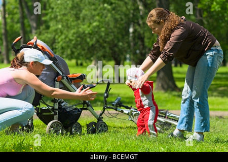 Zwei Frauen helfen, Kleinkind, erste Schritte im Park zu tun. Grünen Rasen und Laub rund um sie. Es ist ein Sommer. Kinderwagen und Fahrrad Stockfoto