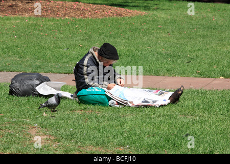 ein Obdachloser ältere Stadtstreicherin auf dem Rasen in der Sonne sitzen und lesen eine Zeitung Stockfoto