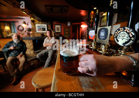 ZWEI MÄNNER SITZEN AM OFFENEN KAMIN IN EINEM TRADITIONELLEN PUB, WIE DIE WIRTIN EINEN PINT BIER AN DER BAR STELLT GLOUCESTERSHIRE ENGLAND UK Stockfoto