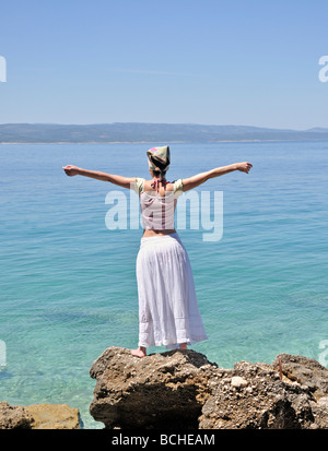 Junge Frau, die auf Felsen von Ozean erhobenen Armen Stockfoto