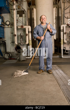 Männlichen kaukasischen im blauen Hausmeister/Hausmeister Overall in Werkseinstellung einschließlich Kessel, Rohre, Ventile.  Er hält Mop. Stockfoto