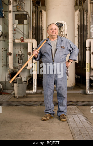 Männlichen kaukasischen im blauen Hausmeister/Hausmeister Overall in Werkseinstellung einschließlich Kessel, Rohre, Ventile.  Er hält Mop. Stockfoto