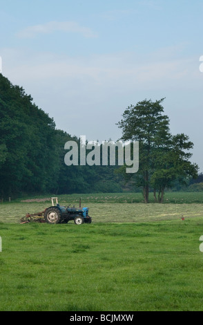 Alten Traktor mit Mähen Gerät stehen verlassen in einem Feld mit einem Wald im Hintergrund Stockfoto