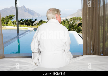 Rückansicht des Mann mittleren Alters sitzen auf dem Bett mit Blick zum pool Stockfoto