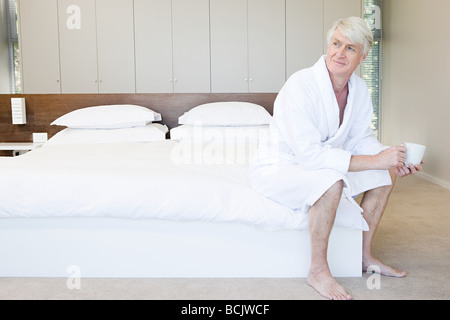 Applying Mann im Bademantel sitzen auf dem Bett mit Tasse Kaffee Stockfoto