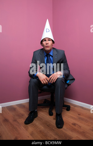 Ein Geschäftsmann saß in der Ecke des Raumes einen Hut Dunce s mit einem dummen Ausdruck auf seinem Gesicht Stockfoto