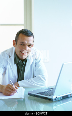 Arzt sitzen mit Laptop-Computer, schreiben mit Stift auf Papier, smiliing Stockfoto