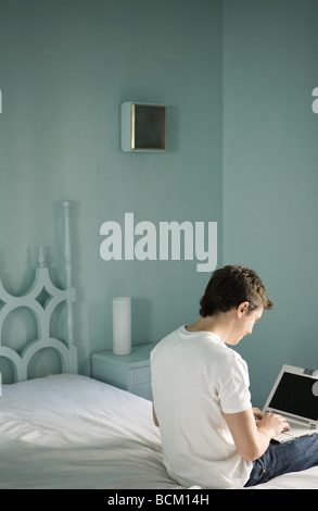 Junger Mann sitzt auf Seite des Bettes, tippen auf laptop Stockfoto