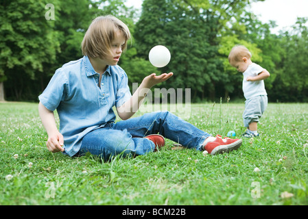 Mädchen mit Down-Syndrom auf dem Boden sitzend wirft Ball in der Luft, Baby Junge stand im Hintergrund Stockfoto