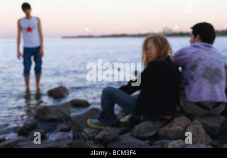 Drei junge Freunde am felsigen Ufer, eine stehend im Wasser, zwei sitzen auf den Felsen Stockfoto