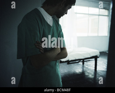 Junger Mann in Scrubs stehend mit Arme verschränkt, Rückblick auf Zimmer mit leeren Bett Stockfoto