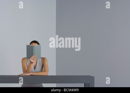 Frau sitzt am Tisch lesen, Gesicht verdeckt durch Buch Stockfoto