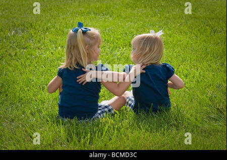 Zwillingsmädchen sitzen auf der Wiese mit Armen auf einander beide gekleidet in blau Stockfoto