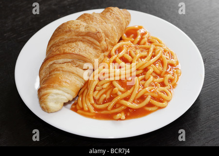 Alternative Frühstück Idee eines frisch gebackenen Croissant mit der Dose Spaghetti in Tomatensoße vor einem schwarzen Hintergrund mit Keine Personen Stockfoto