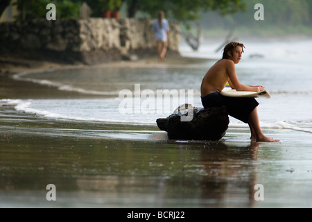Ein junger Surfer sitzt auf einem Baumstamm auf Playa Tamarindo in Guanacaste, Costa Rica, hält sein Board und ruhen, während die Wellen warten. Stockfoto