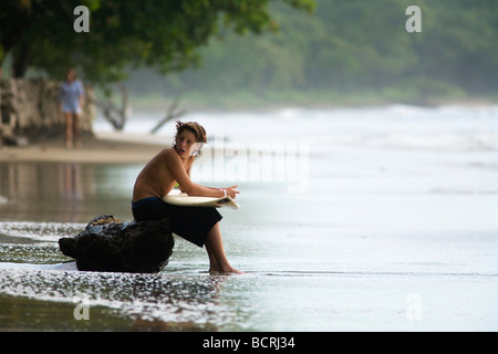 Ein junger Surfer sitzt auf einem Baumstamm auf Playa Tamarindo in Guanacaste, Costa Rica, hält sein Board und ruhen, während die Wellen warten. Stockfoto