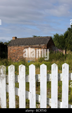 Eine verfallene Scheune auf einem Stück Ödland hat Brettern vernagelt, um Eintrag zu beschränken und sitzt hinter einem alten weißen Zaun Stockfoto