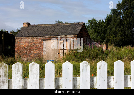 Eine verfallene Scheune auf einem Stück Ödland hat Brettern vernagelt, um Eintrag zu beschränken und sitzt hinter einem alten weißen Zaun Stockfoto