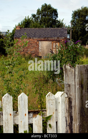Eine verfallene Scheune auf einem Stück Ödland hat Brettern vernagelt, um Eintrag zu beschränken und sitzt hinter einem alten weißen Zaun Stockfoto
