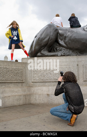 Ein junger Mann fotografiert eine hübsche blonde Mädchen in einem gelben Mantel und rote Kniestrümpfe Socken von den Löwen auf dem Trafalgar Square posiert Stockfoto