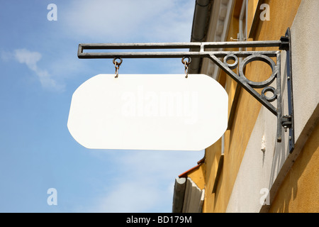 Leere, weiße Ladenschild hängen in einer schmiedeeisernen Halterung auf einer gelben Wand vor einem blauen Himmel. Stockfoto