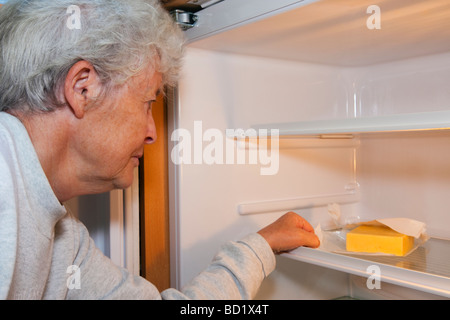 Ältere Frau Rentner OAP mit betroffenen Ausdruck suchen in einem fast leeren Kühlschrank mit nur einem Stück Käse aufgrund von Sparmaßnahmen. Großbritannien Großbritannien Stockfoto