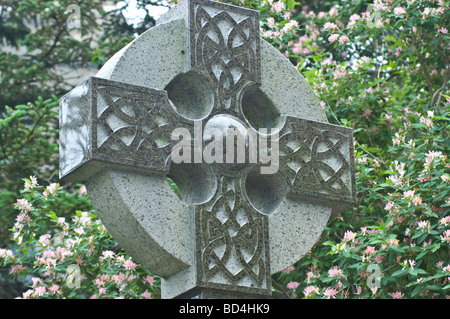Aufwändig geschnitzte Grabstatue aus Stein des Keltischen Kreuzes auf einem Friedhof in Halifax, Nova Scotia, Kanada, umgeben von üppigem Grün und rosa Blumen. Stockfoto
