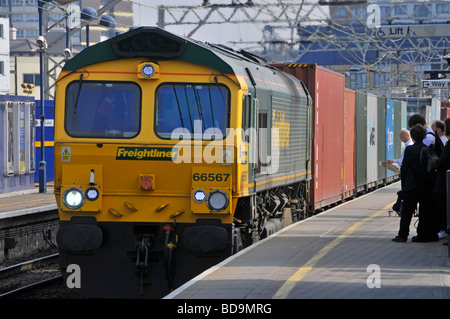 Stratford East London Freightliner Lokomotive 66567 mit Schiffscontainern Güterzug, der auf Bahnsteig England UK wartende Passagiere passiert Stockfoto