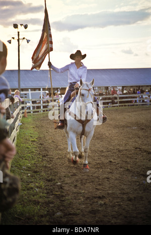 Junge Frau auf weißem Pferd trägt amerikanische Fahne bei der Eröffnungsfeier der Kirmes rodeo Stockfoto